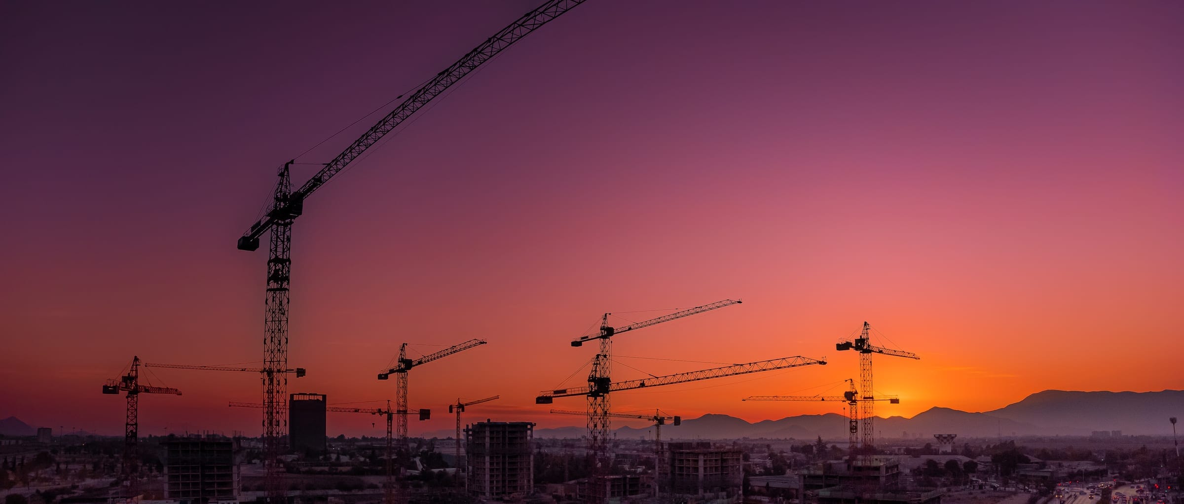 Tower cranes over buildings under construction, silhouetted against a sunset sky