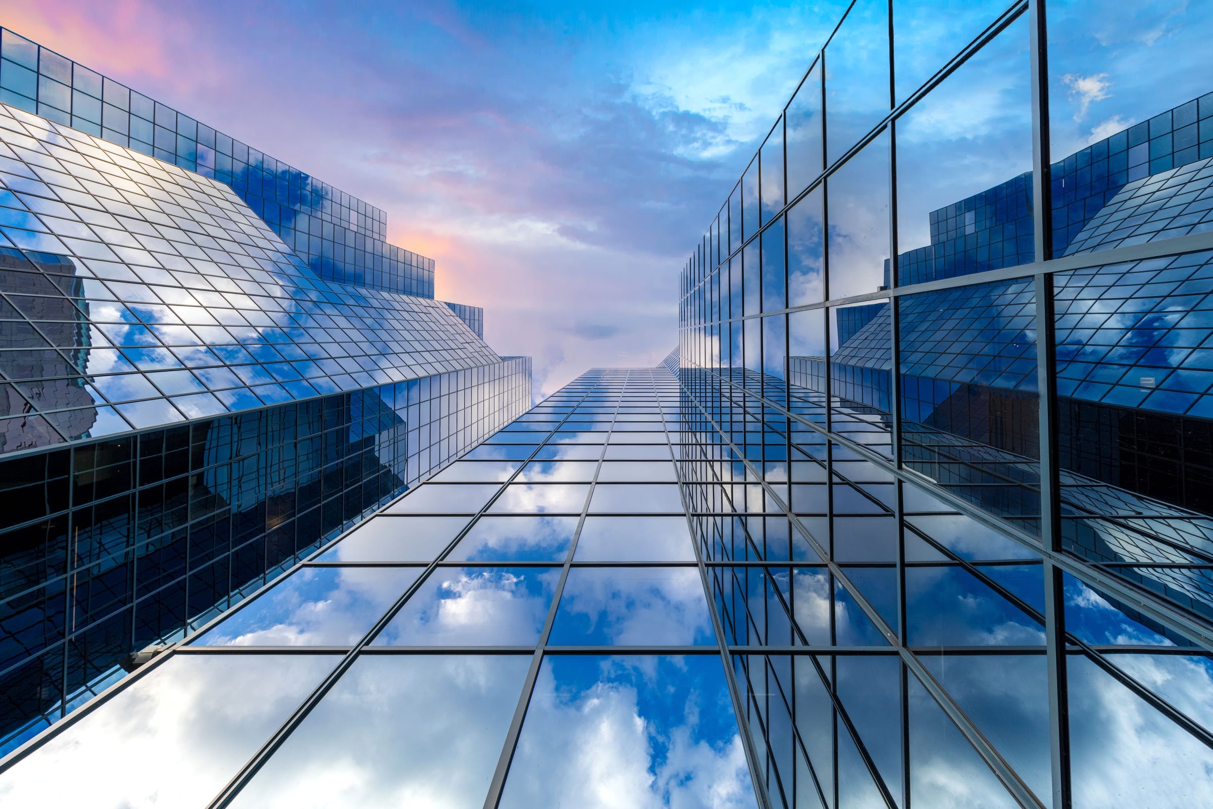 Looking up between glass office towers that reflect a blue sky and clouds