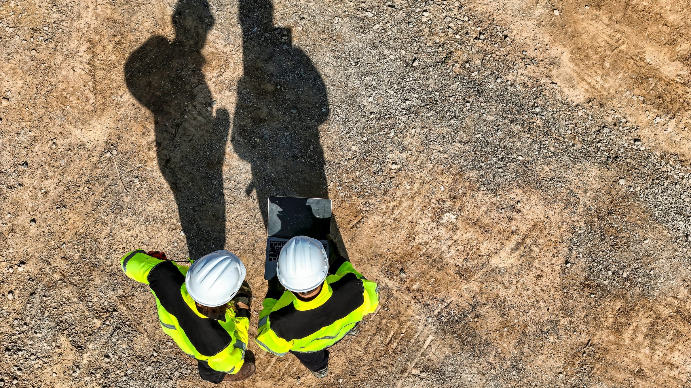Aerial view of two site workers in hard hats and high-visibility jackets reviewing a laptop on a construction site