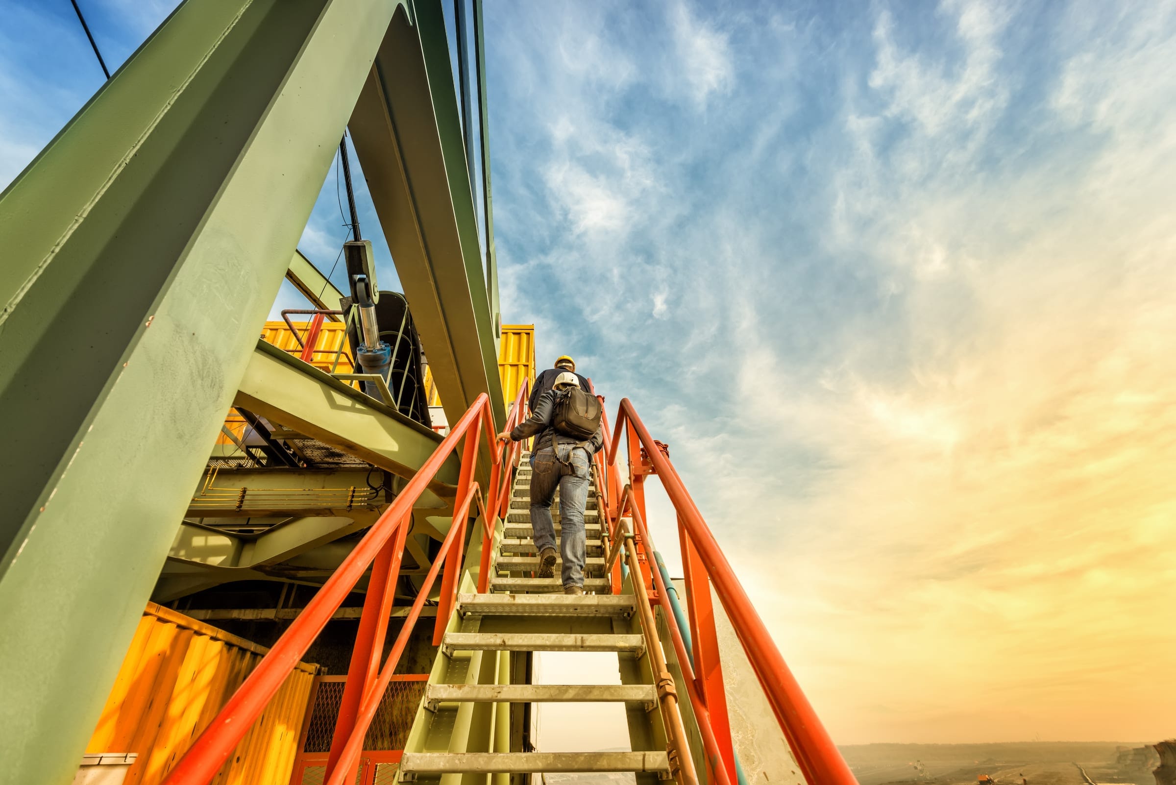 Two workers in hard hats climbing an orange-railed steel stairway on an industrial structure under a bright sky