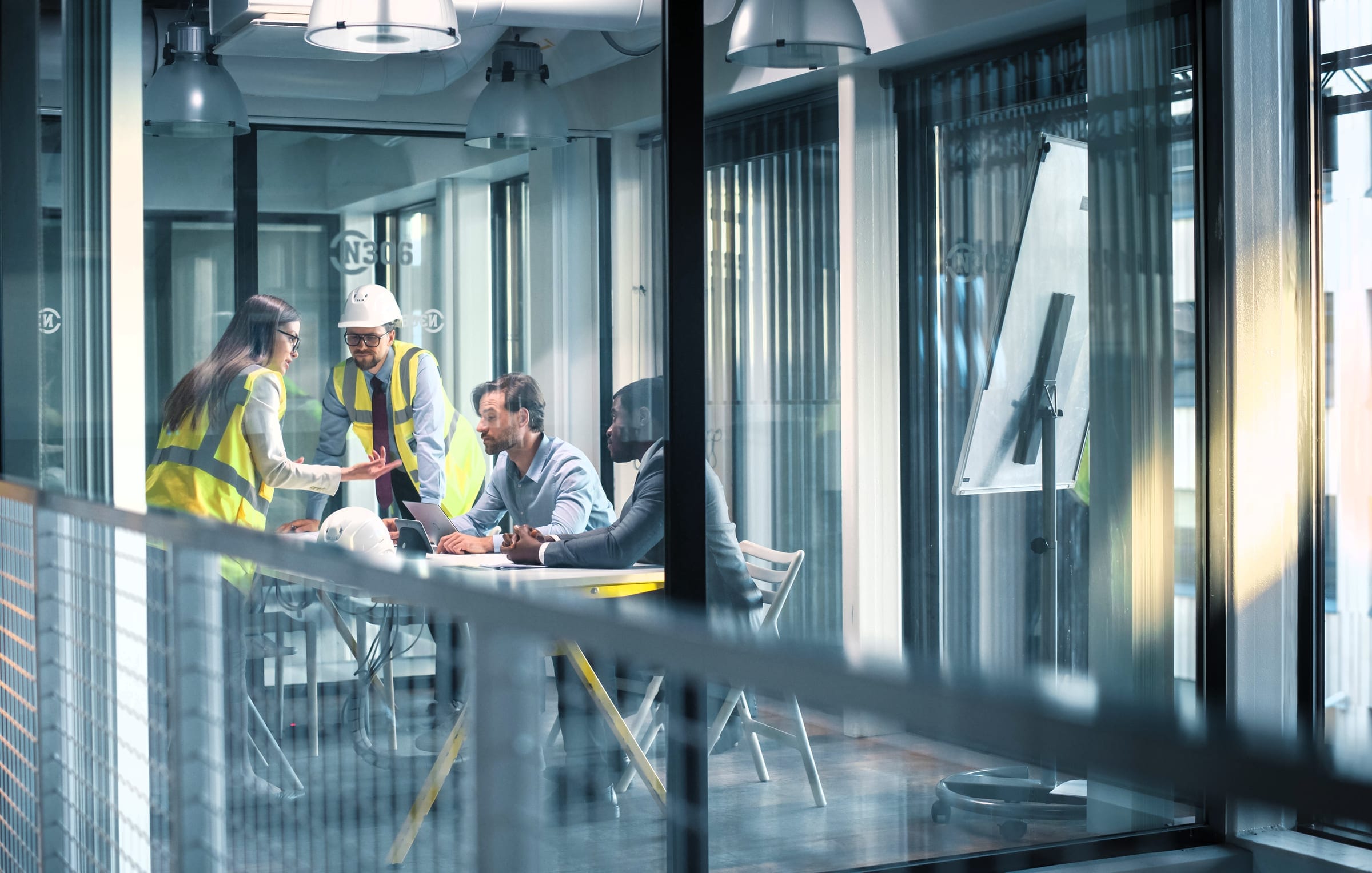 Four colleagues, two in high-visibility vests, discussing plans around a table in a glass-walled meeting room